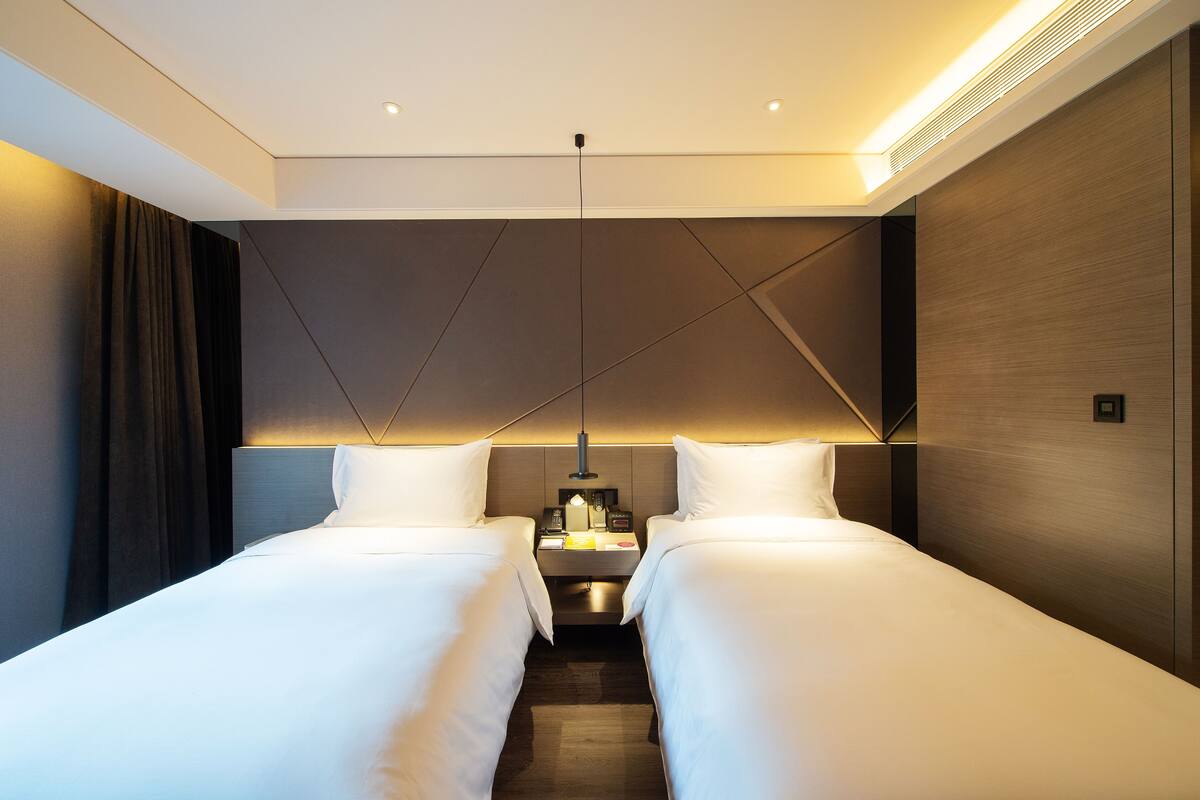 A modern hotel room with two single beds, a bedside table with a lamp, and a headboard with geometric patterns.