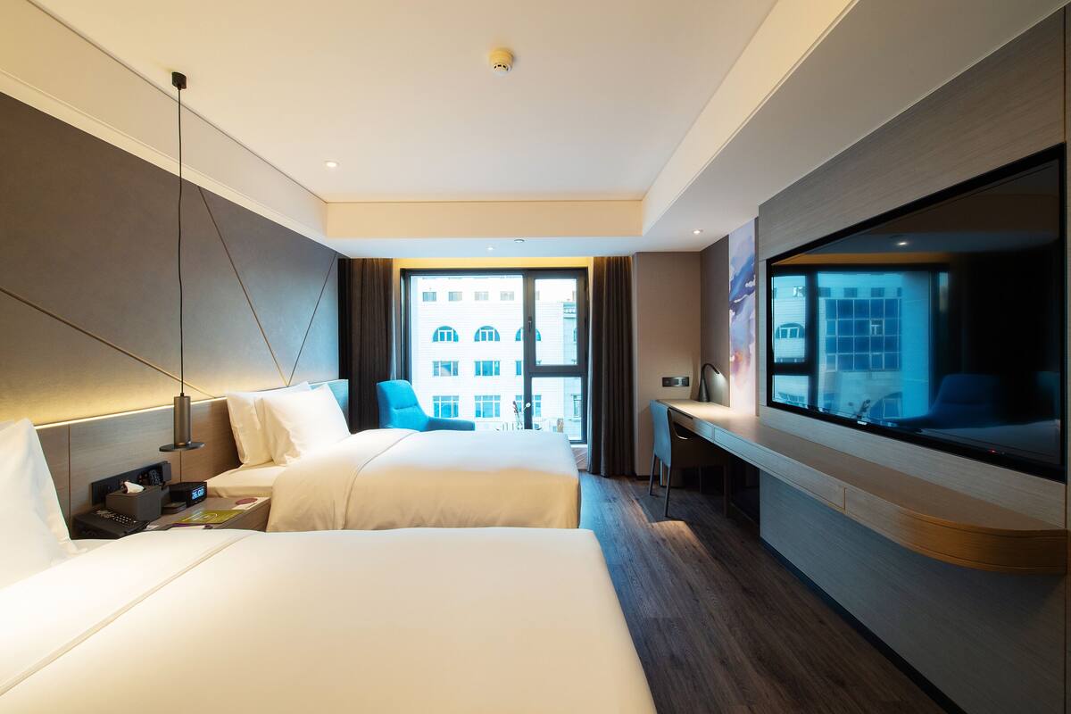 A modern hotel room with two beds, a desk, and a large window offering a city view.