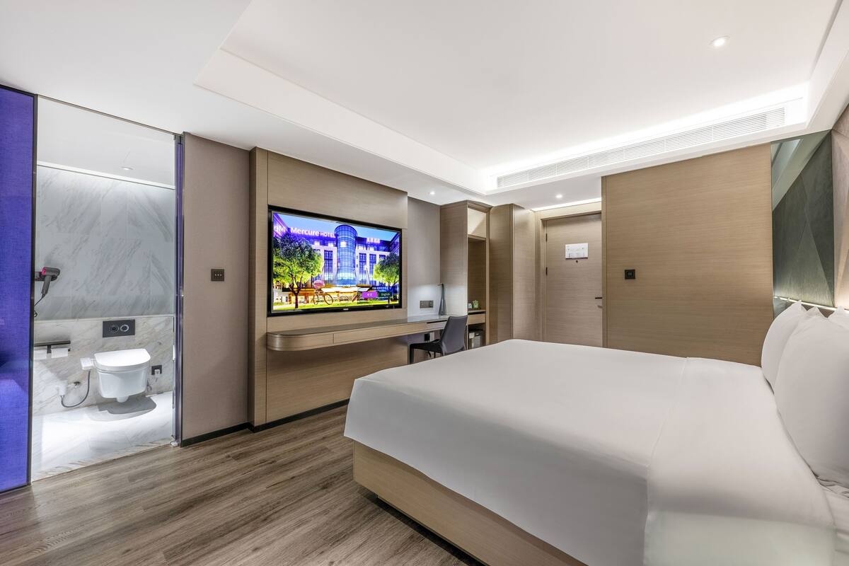 A modern hotel room with a large bed, a television, a desk, and a bathroom.