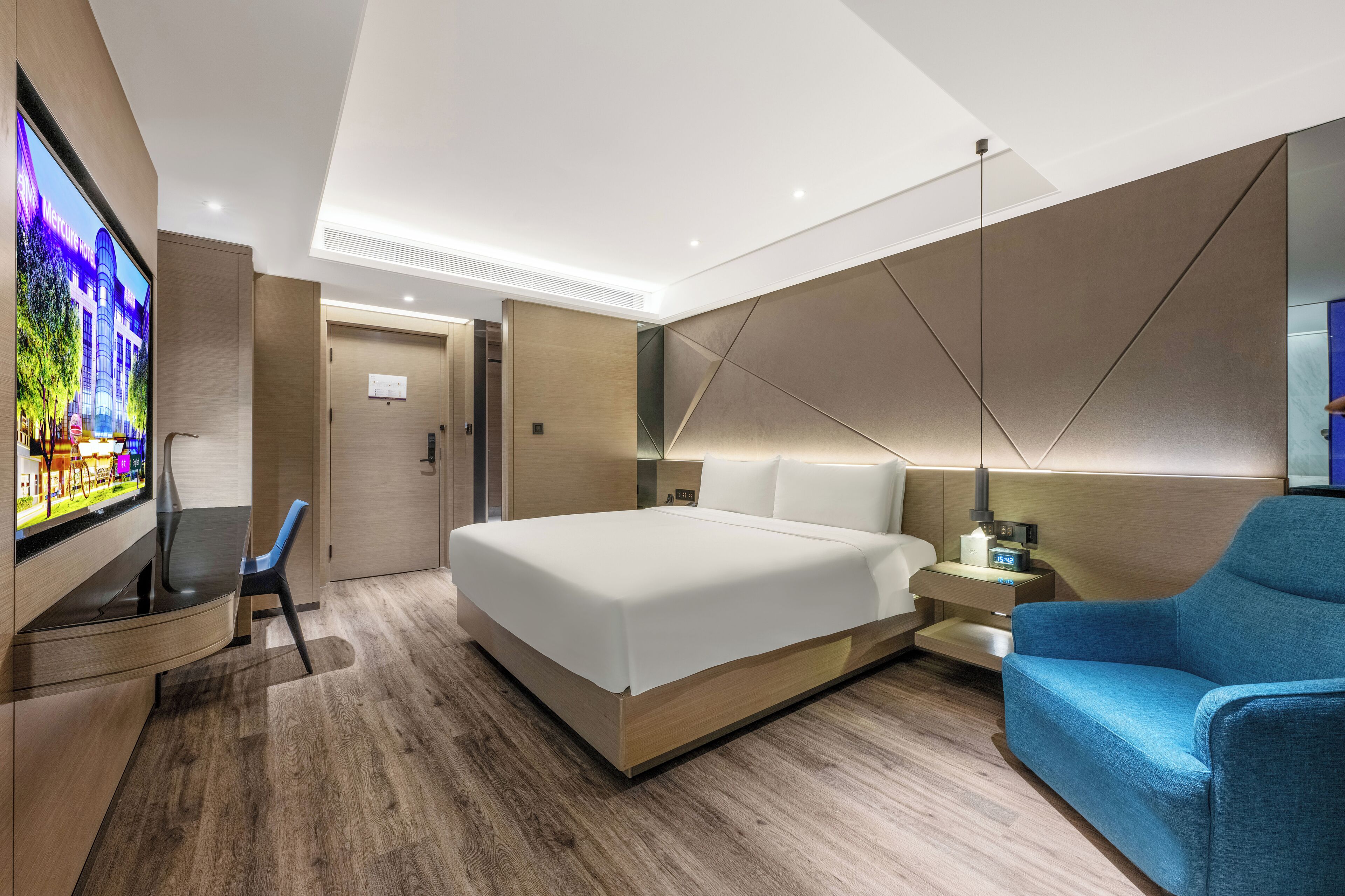 A modern hotel room with a large bed, a TV, a desk, and a blue armchair.