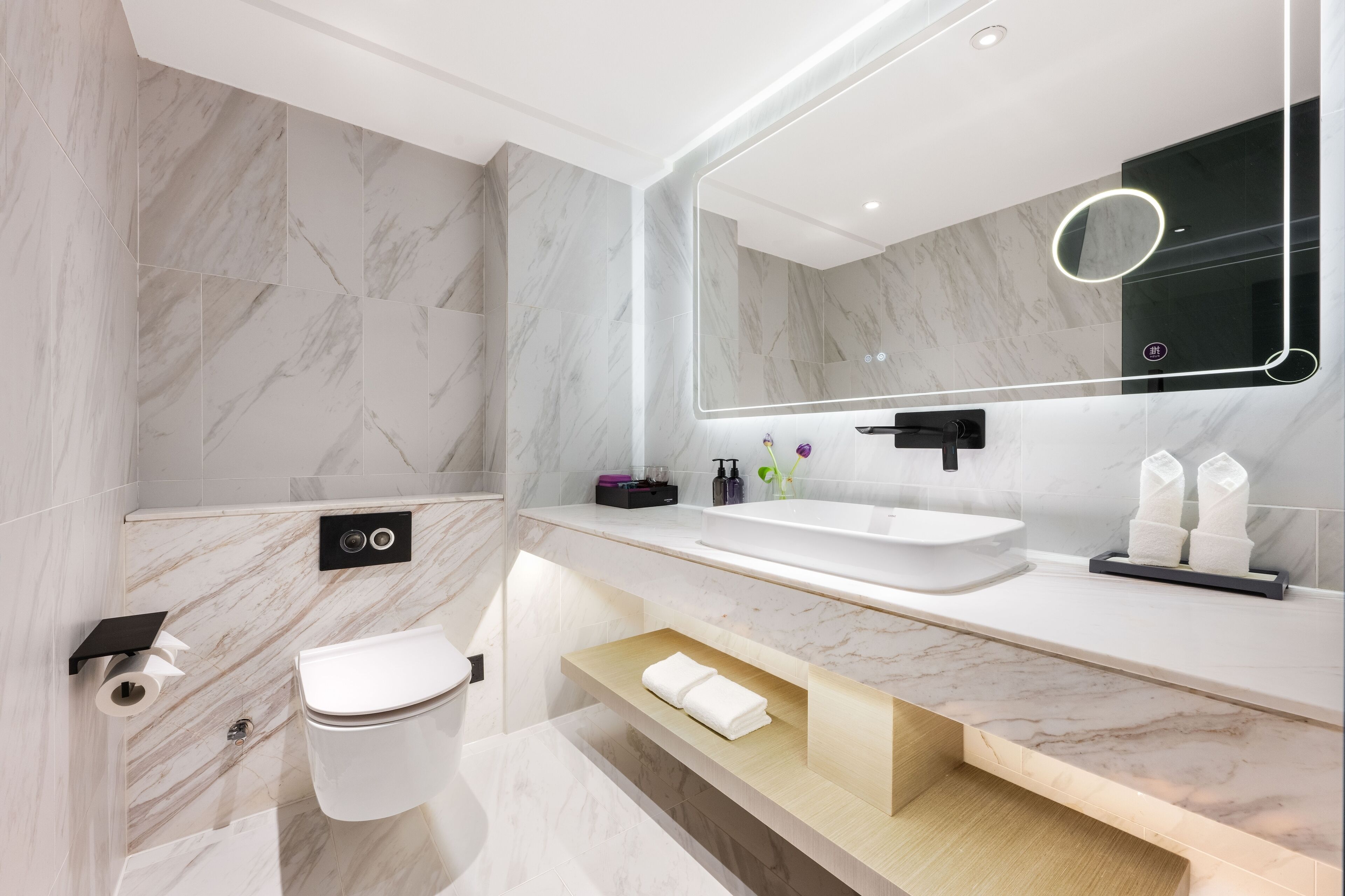 A modern bathroom with a marble sink, a toilet, a mirror with integrated lighting, and a towel rack.