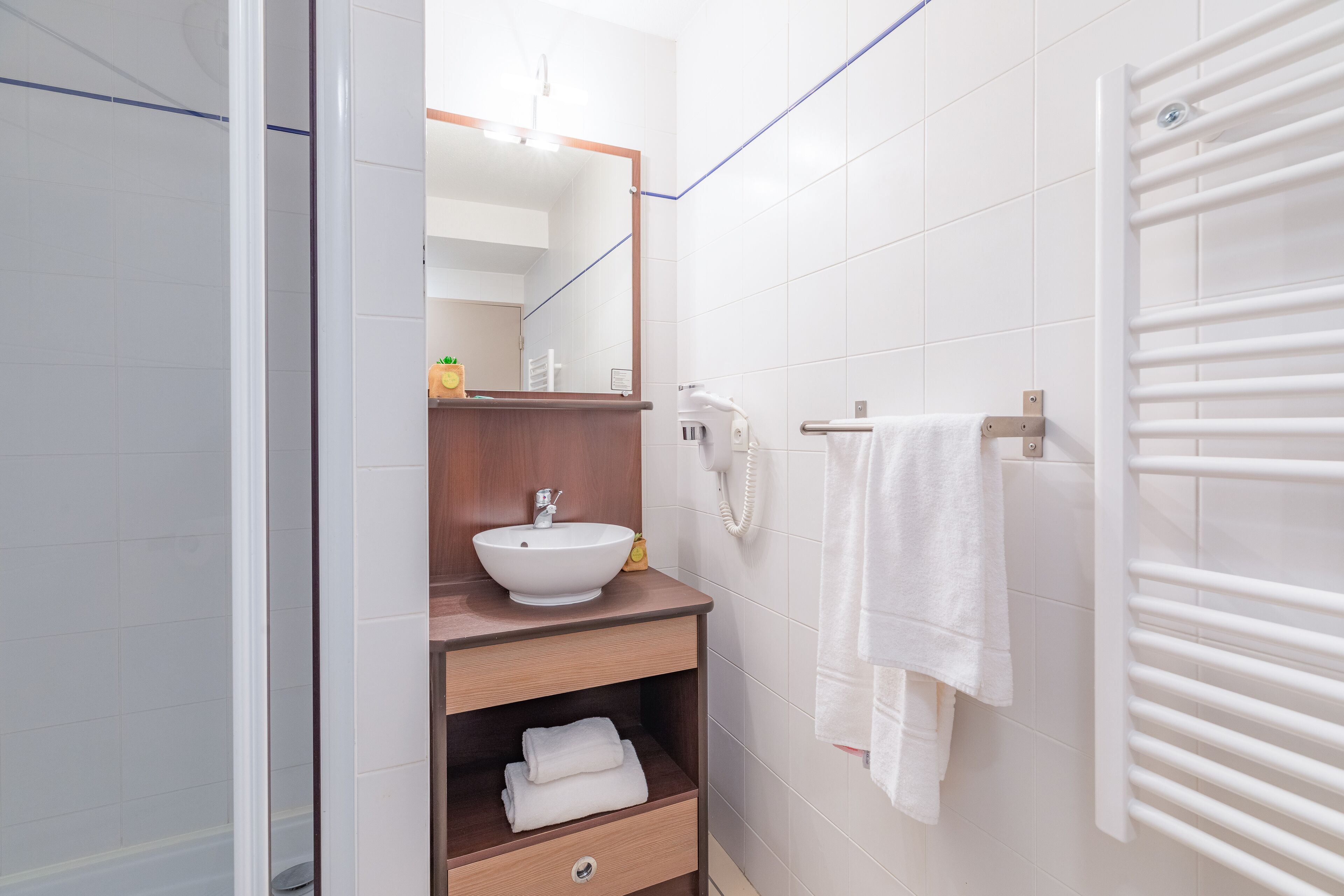 A modern bathroom with a white sink, wooden vanity, a mirror, a towel rack, and a shower area.