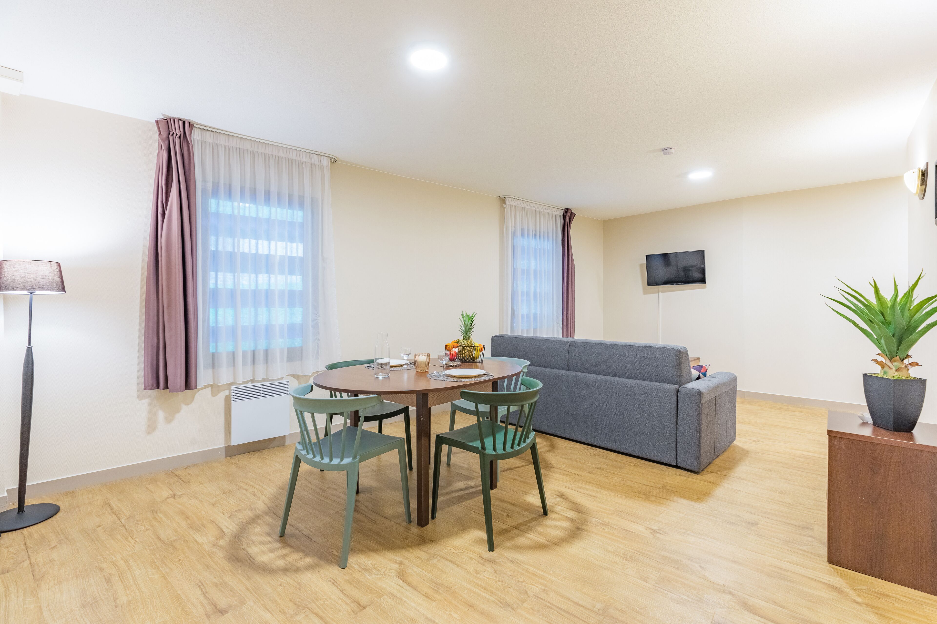 A modern living room with a dining table, chairs, a sofa, a TV mounted on the wall, and a potted plant.