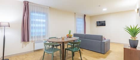 A modern living room with a dining table, chairs, a sofa, a TV mounted on the wall, and a potted plant.