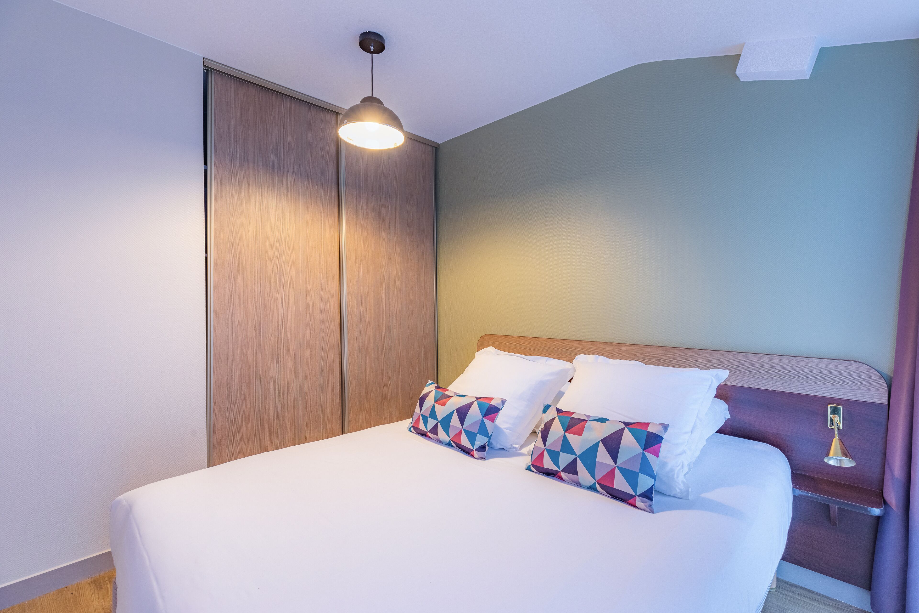 A neatly made bed with white linens and colorful geometric pillows, a wooden headboard, and a bedside table with a lamp.