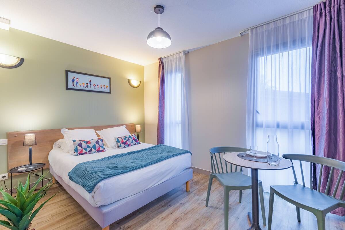 Una habitación de hotel con cama, lámparas de noche, una mesa con sillas y una ventana con cortinas.