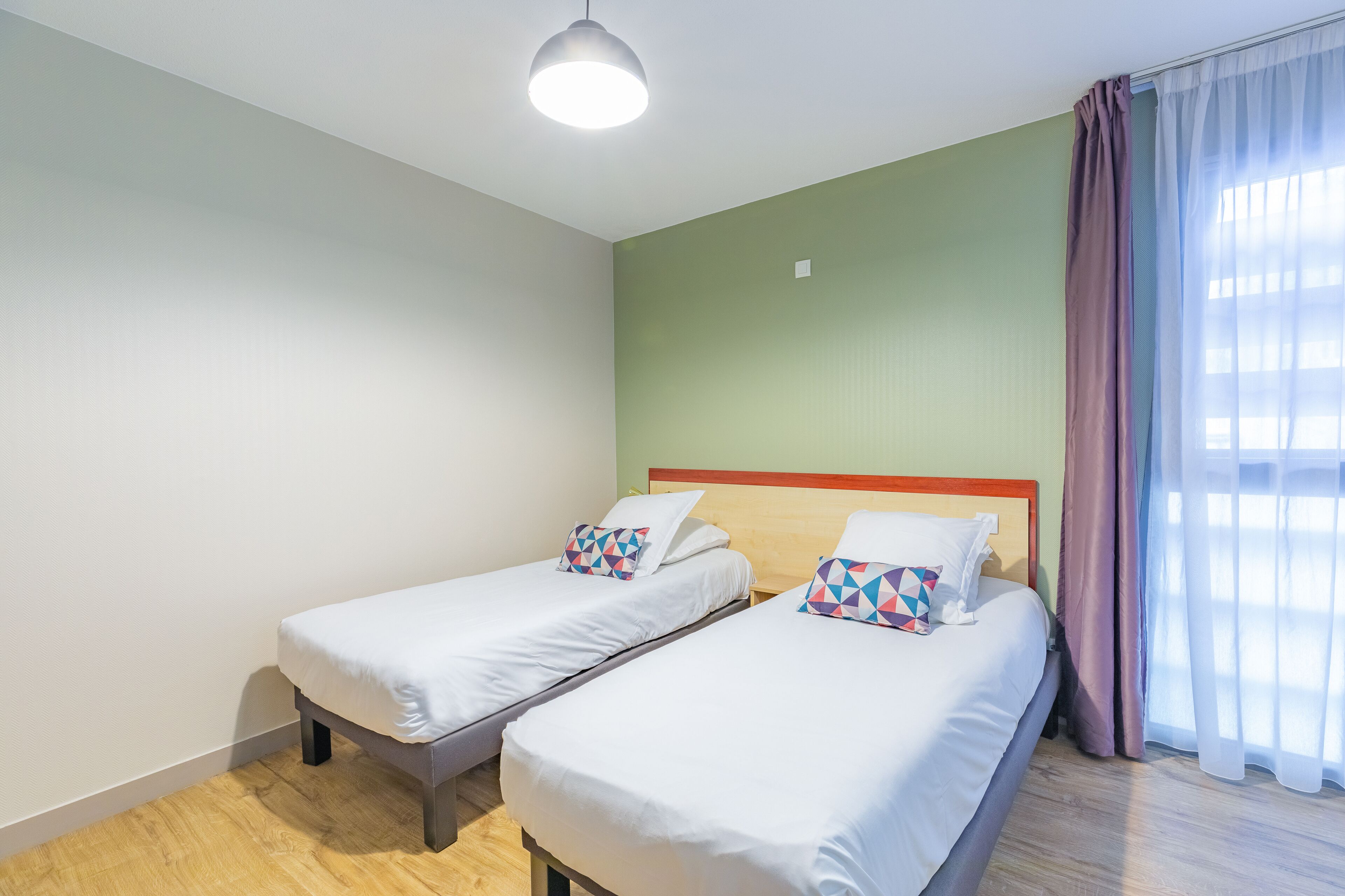 A hotel room with two single beds, a wooden headboard, a green wall, and a large window with sheer curtains.