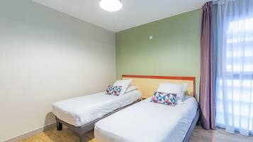 A hotel room with two single beds, a wooden headboard, a green wall, and a large window with sheer curtains.
