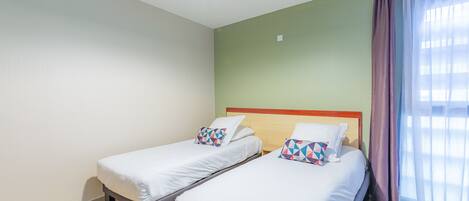A hotel room with two single beds, a wooden headboard, a green wall, and a large window with sheer curtains.