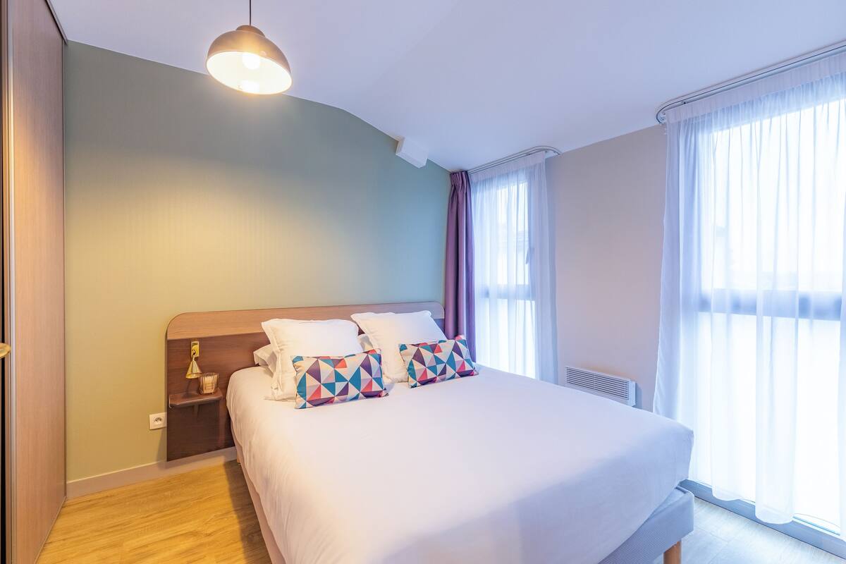 Una cama bien hecha con almohadillas estampadas, una mesita de noche con lámpara y una ventana con cortinas translúcidas.