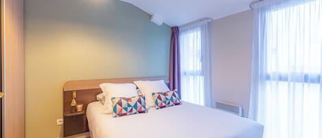 Una cama bien hecha con almohadillas estampadas, una mesita de noche con lámpara y una ventana con cortinas translúcidas.