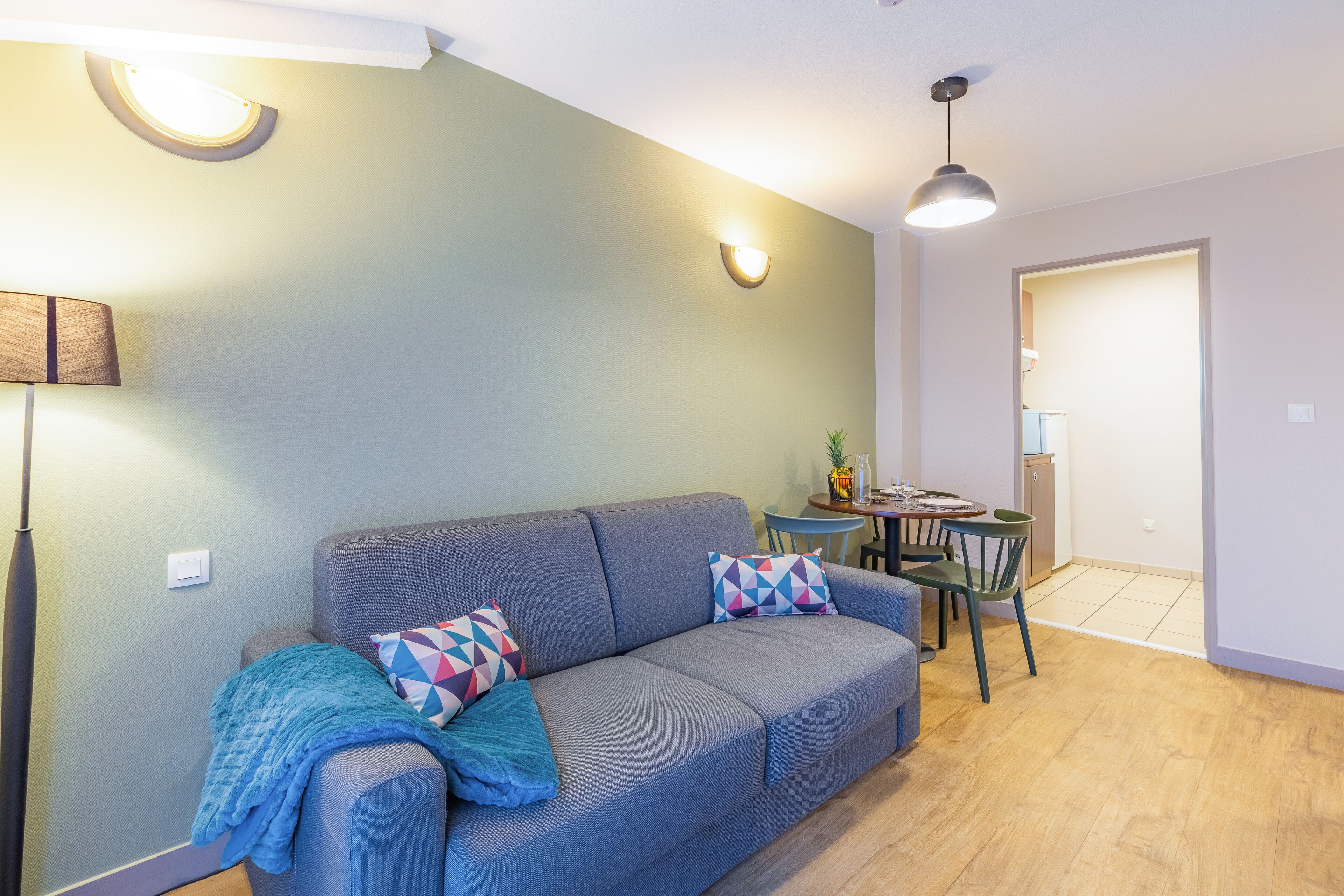 A modern living room with a grey sofa, colorful geometric pillows, a blue throw blanket, a round table with chairs, and a pendant light.