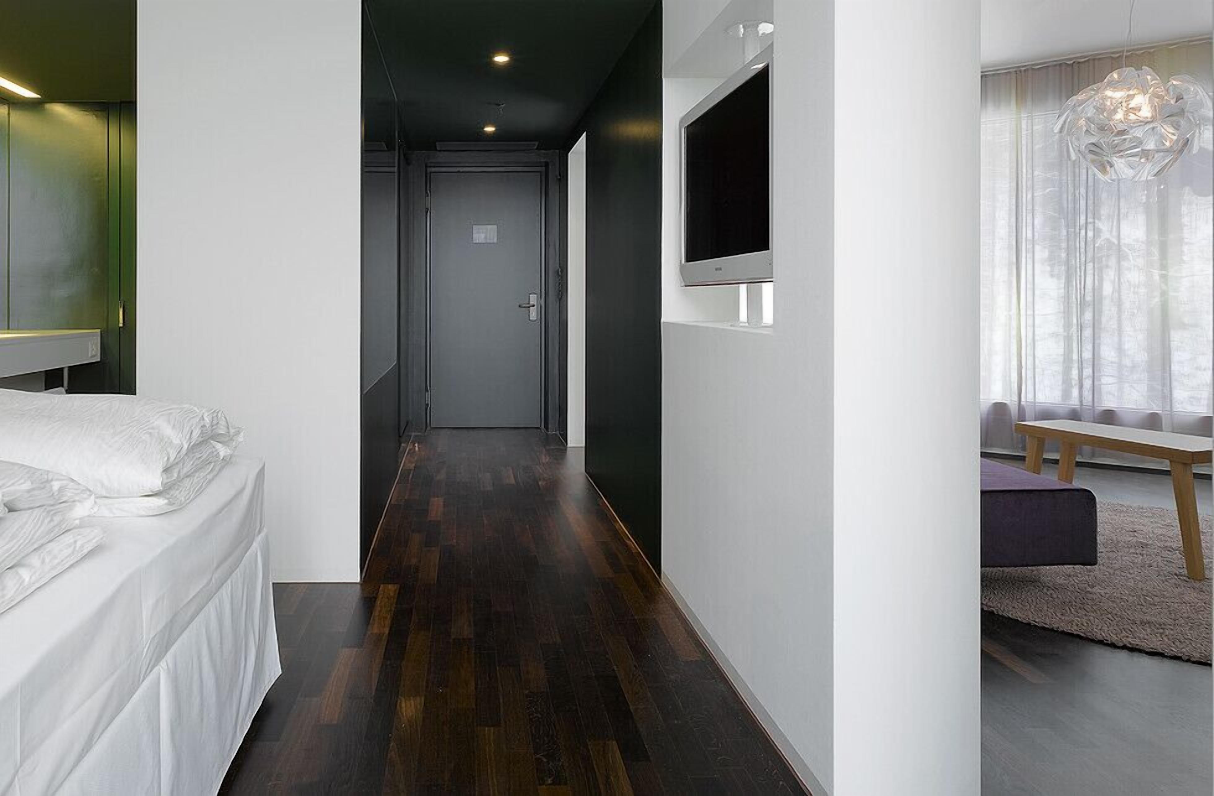 Una habitación de hotel moderna con una cama, un televisor fijado a la pared, suelo de madera y una ventana con cortinas ligeras.