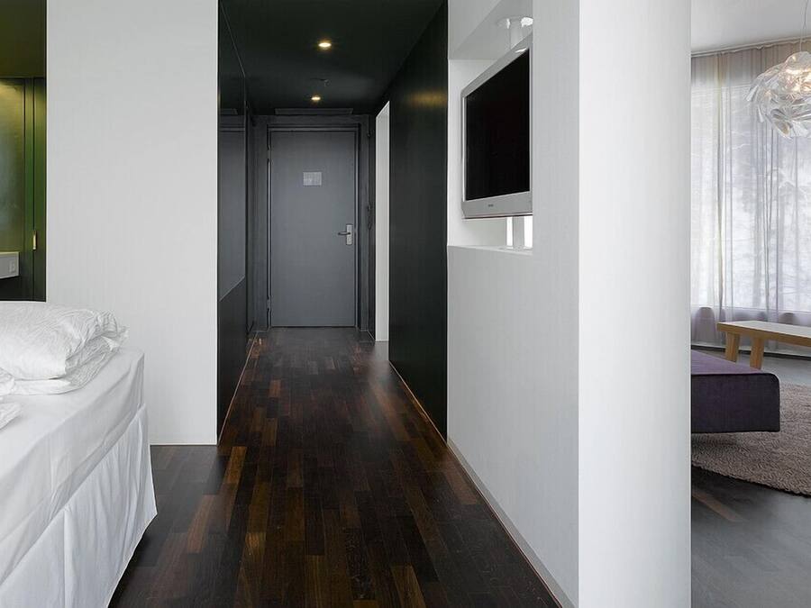 Una habitación de hotel moderna con una cama, un televisor fijado a la pared, suelo de madera y una ventana con cortinas ligeras.