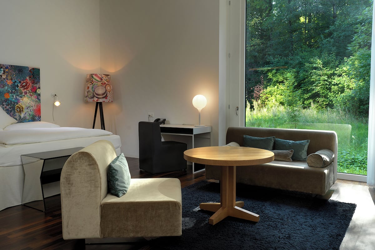 A modern living room with a bed, a sofa, a chair, a round table, and a large window with a view of greenery.