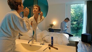 A hotel room with a large mirror, a sink, a bed, a window with a view of trees, and a bedside table with a lamp. - City Garden Hotel (Zug)