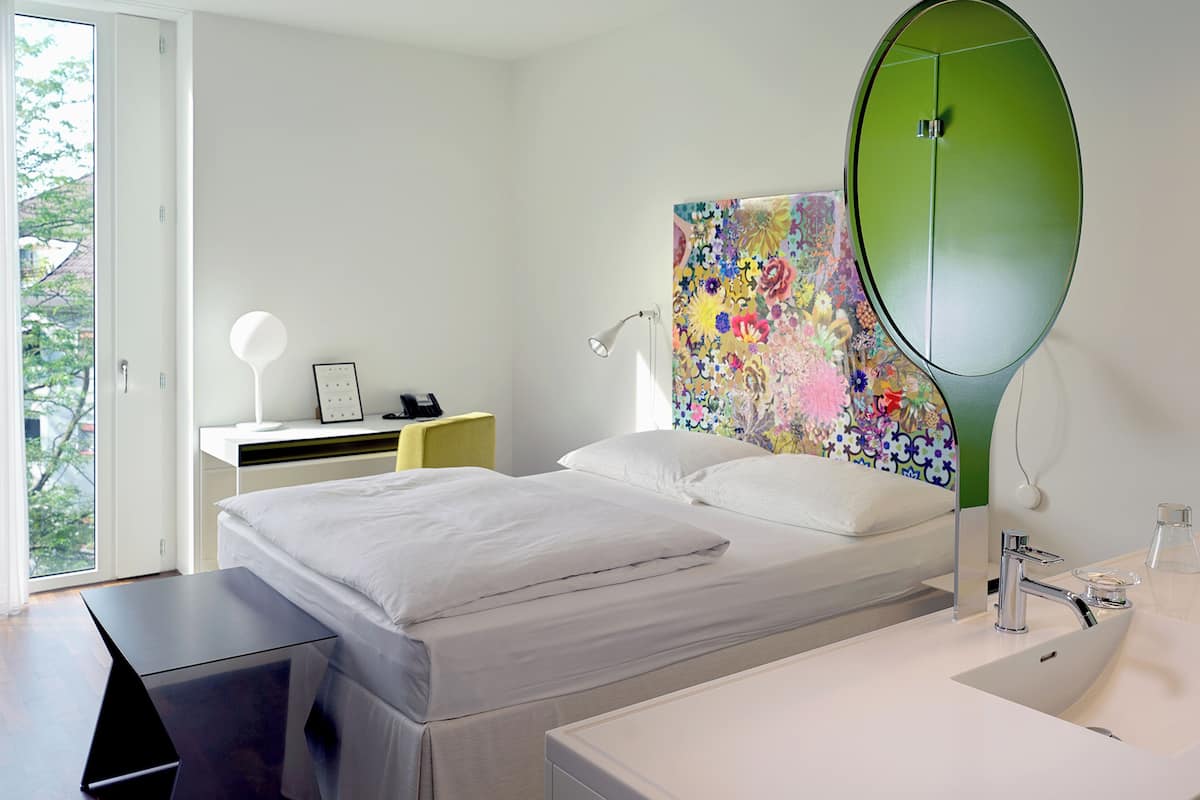 A modern hotel room with a bed, bedside table, lamp, and a large mirror.