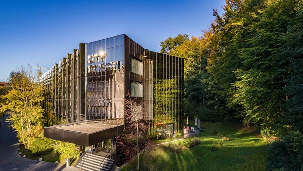 A modern, glass-walled building with a unique cantilevered structure, surrounded by lush greenery. - City Garden Hotel (Zug)