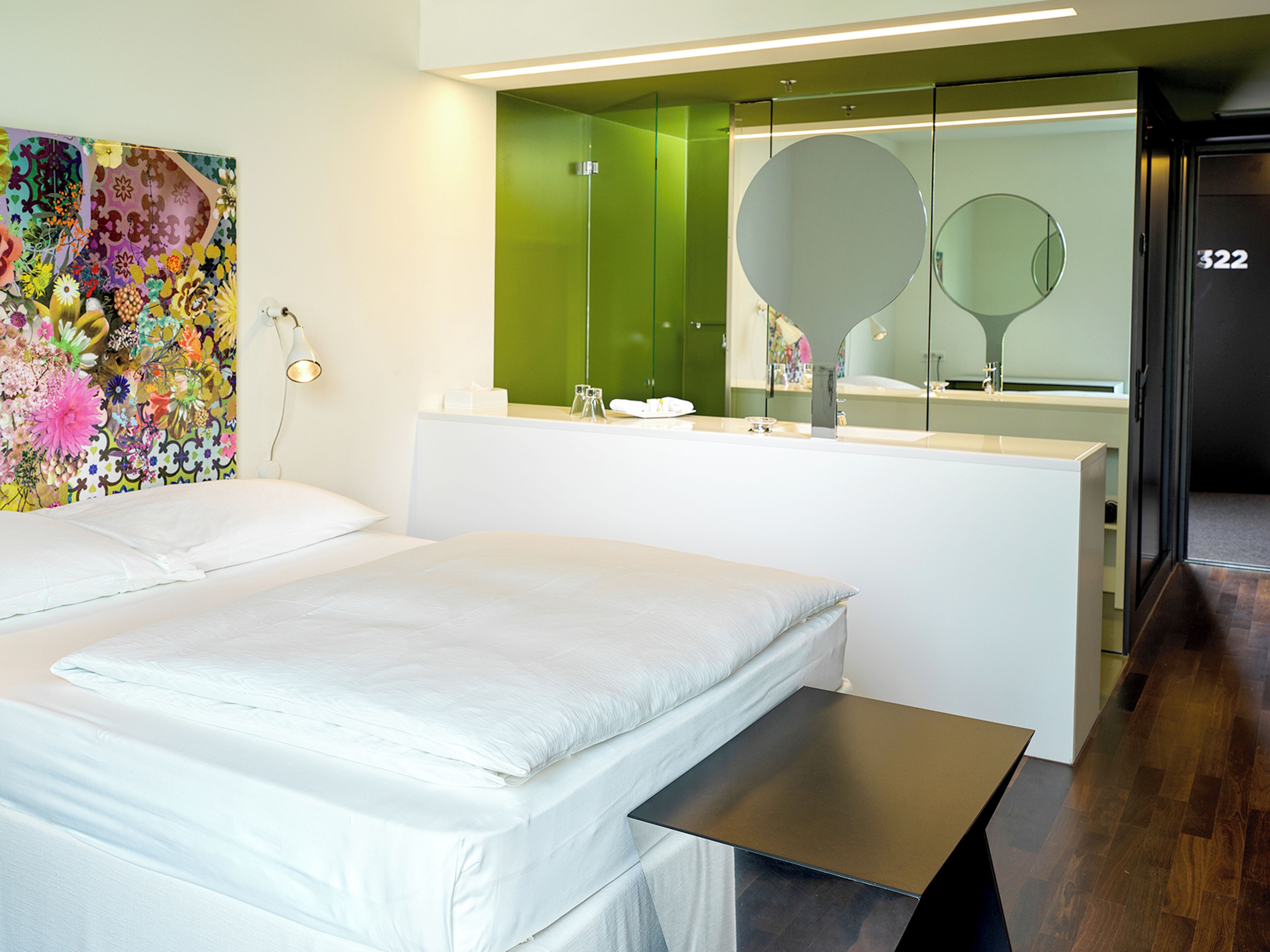 A modern hotel room with a large bed, a bedside table, a mirror, and a green accent wall.
