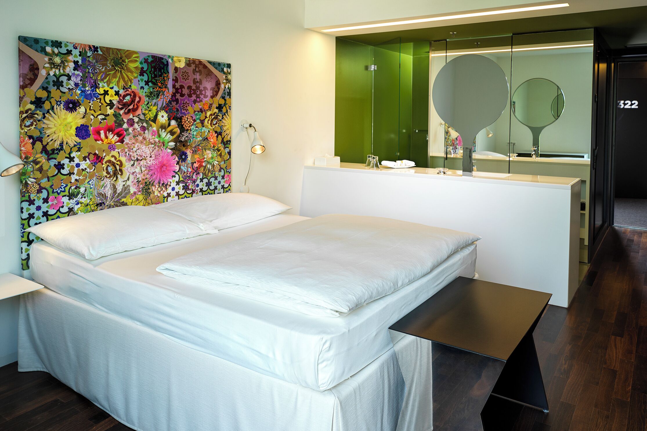 A modern hotel room with a large bed, a bedside table, a mirror, and a colorful floral artwork on the wall.