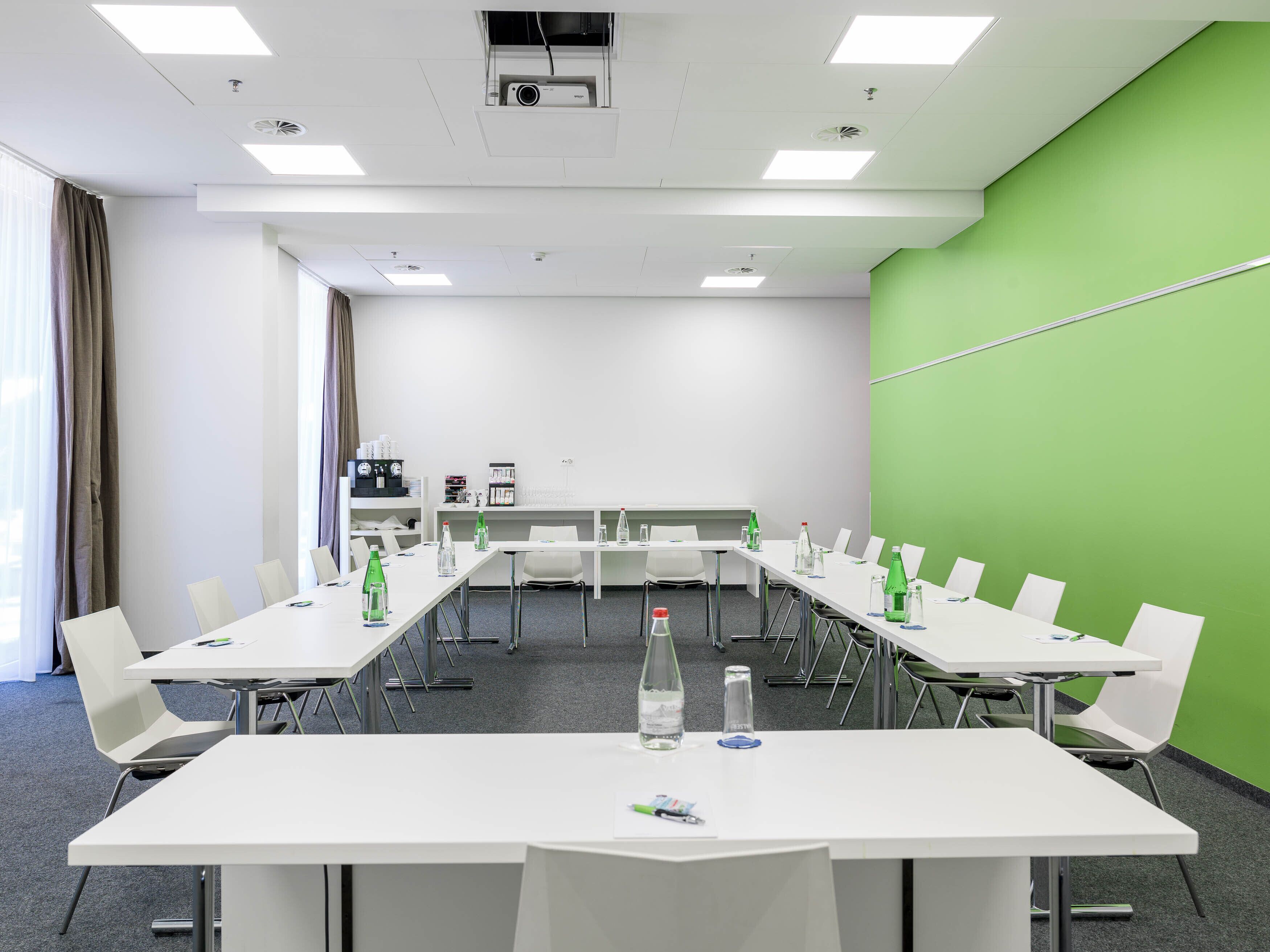 A conference room with white tables and chairs, a projector, and a green accent wall.