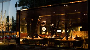 A modern restaurant with a glass facade, interior lighting, and a view of parked cars outside. - City Garden Hotel (Zug)