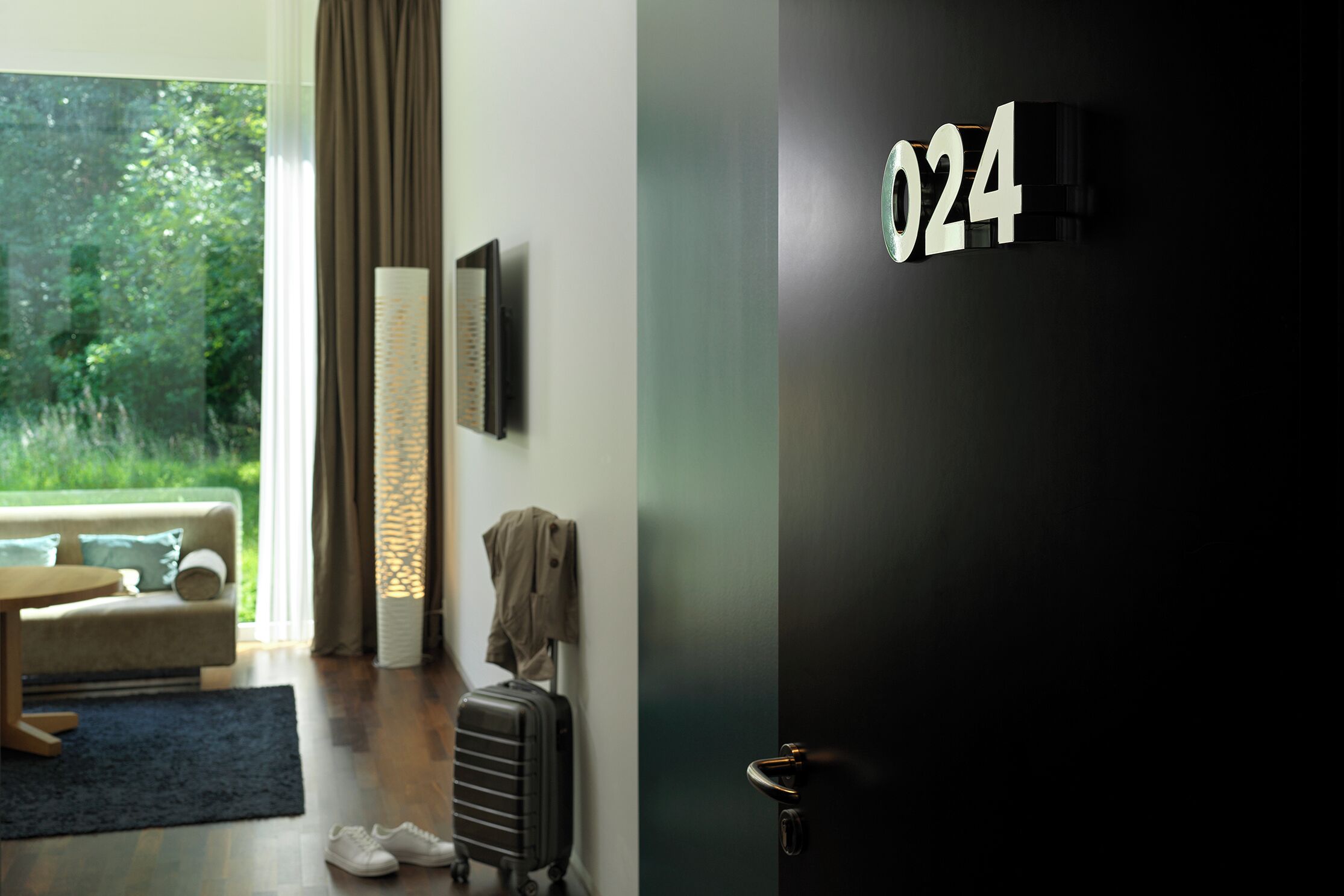 A hotel room with a large window, a sofa, a lamp, a suitcase, and a door with the number 024.