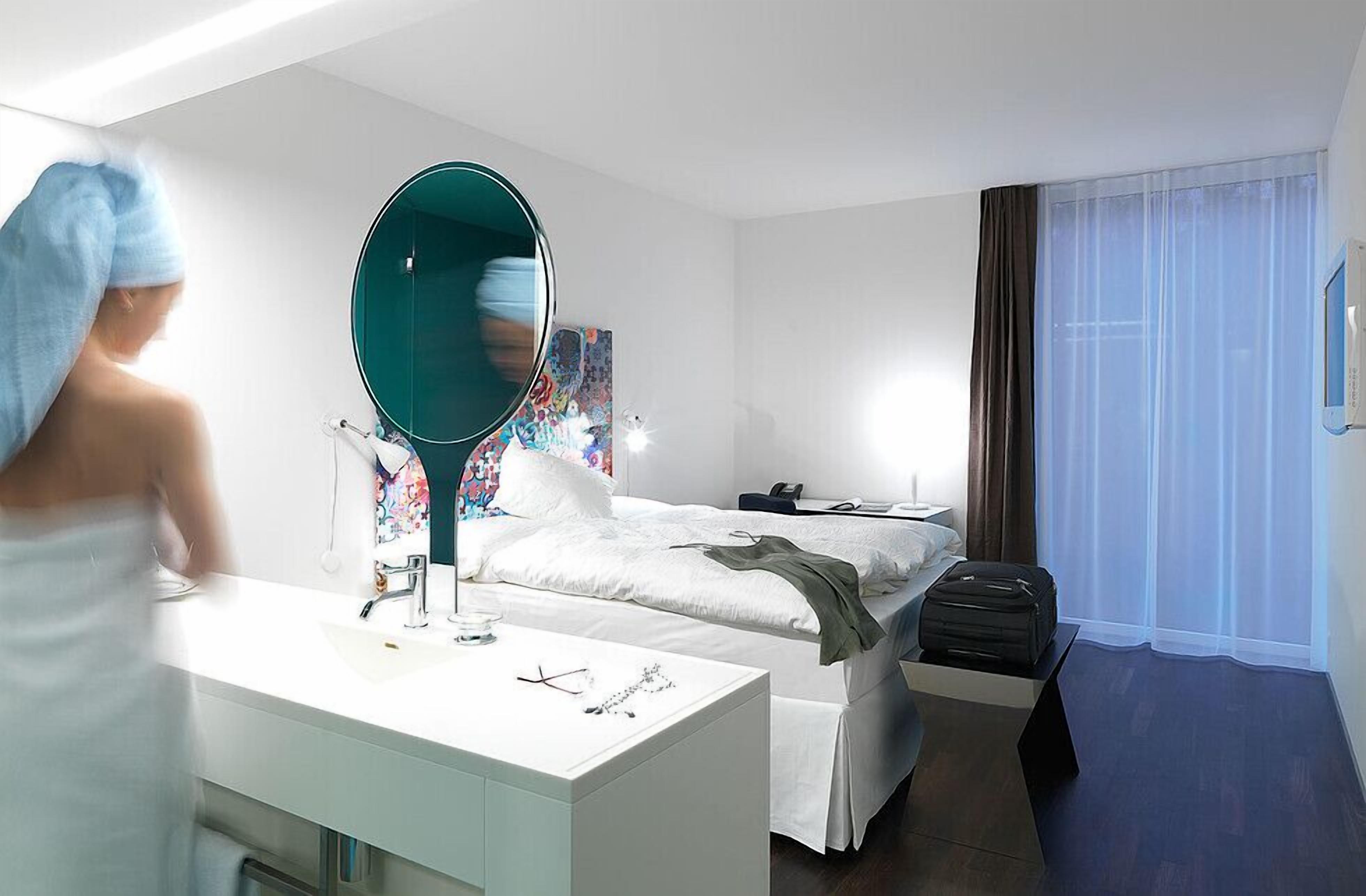A modern hotel room with a large mirror, a bed, a bedside table, a desk, and a window with curtains.