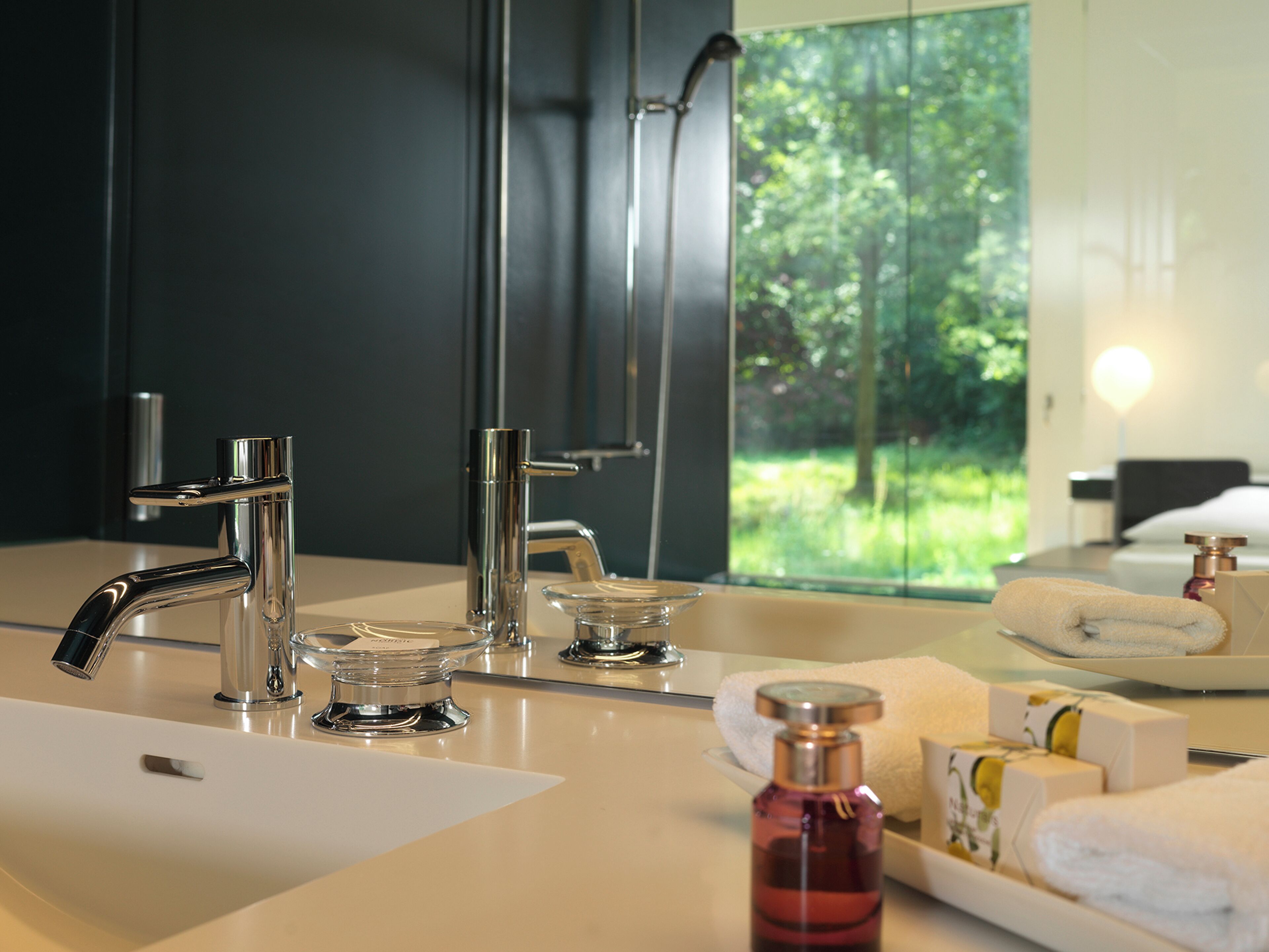 A modern bathroom with a large mirror, two sinks, and a view of greenery outside.