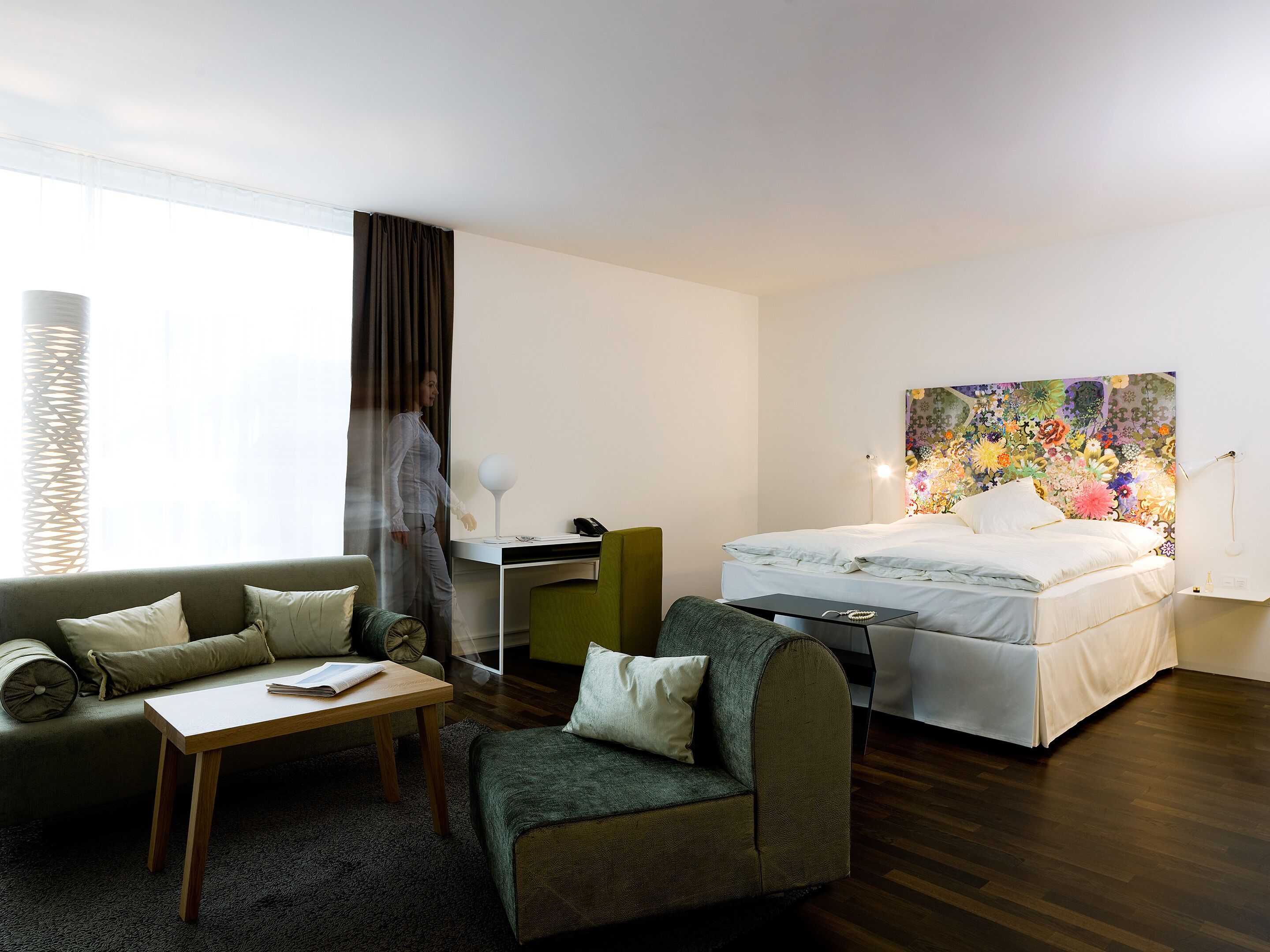 A hotel room with a bed, a desk, a chair, a sofa, and a large window with curtains.