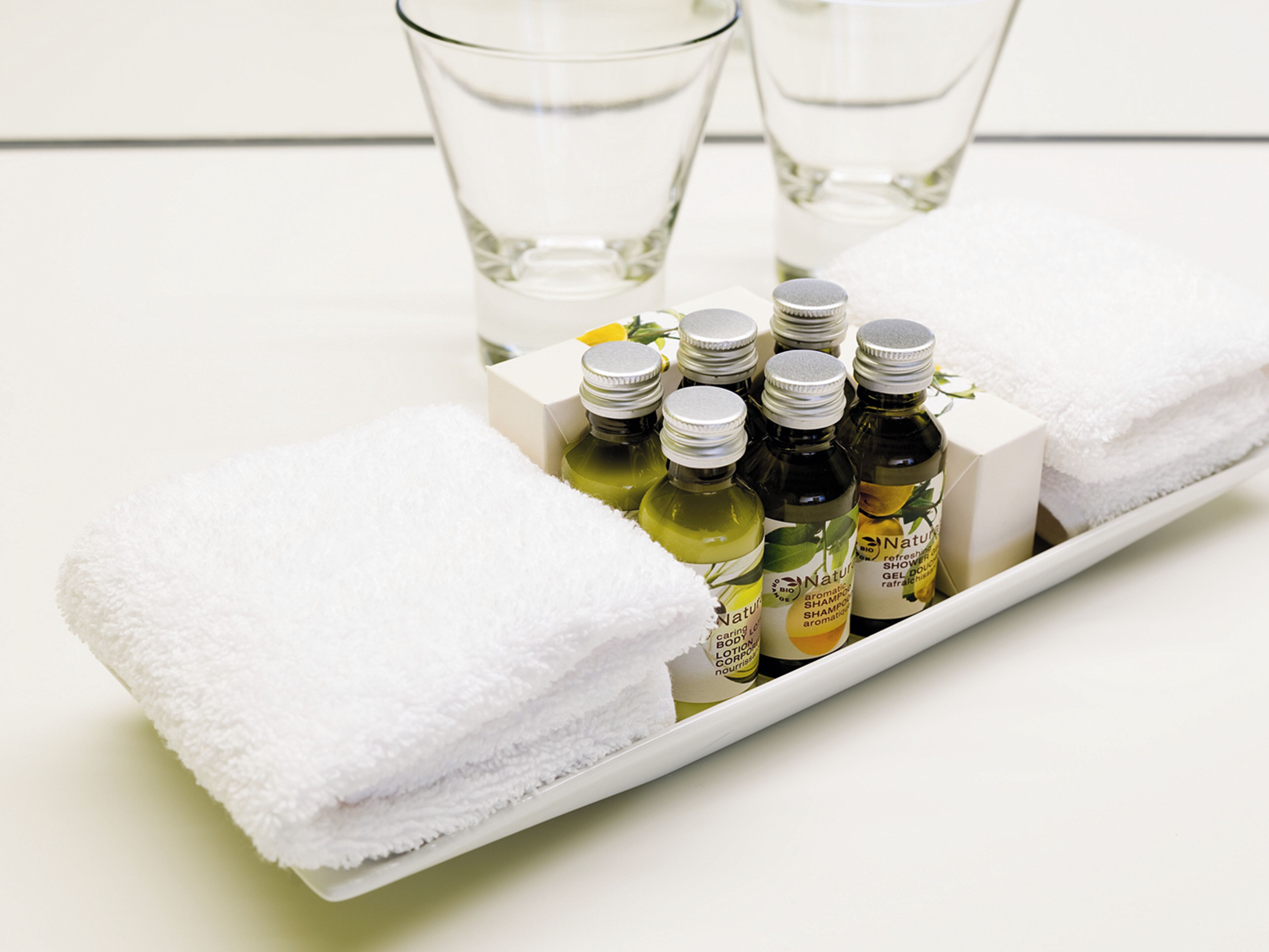 Un plateau avec des flacons de gel douche et de lotion, une serviette blanche et deux verres d’eau.