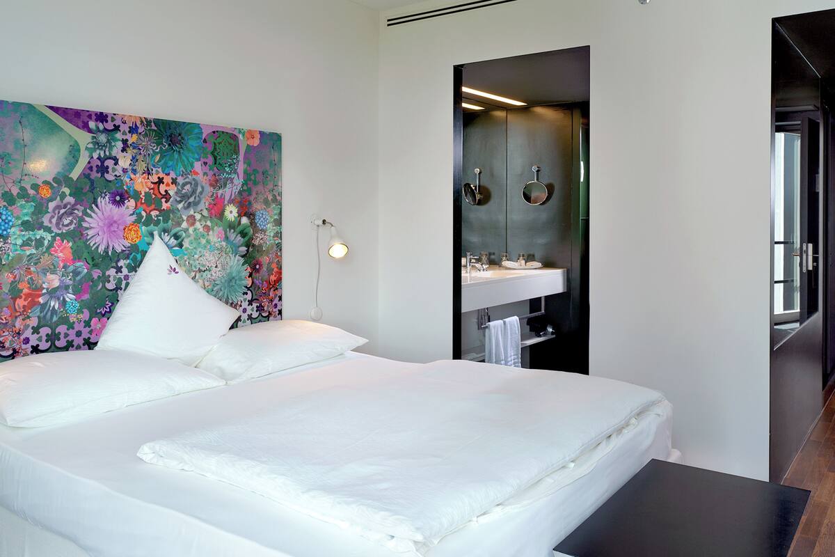 A hotel room with a large bed, a colorful floral headboard, a small bedside table, and a bathroom with a modern sink and mirror.