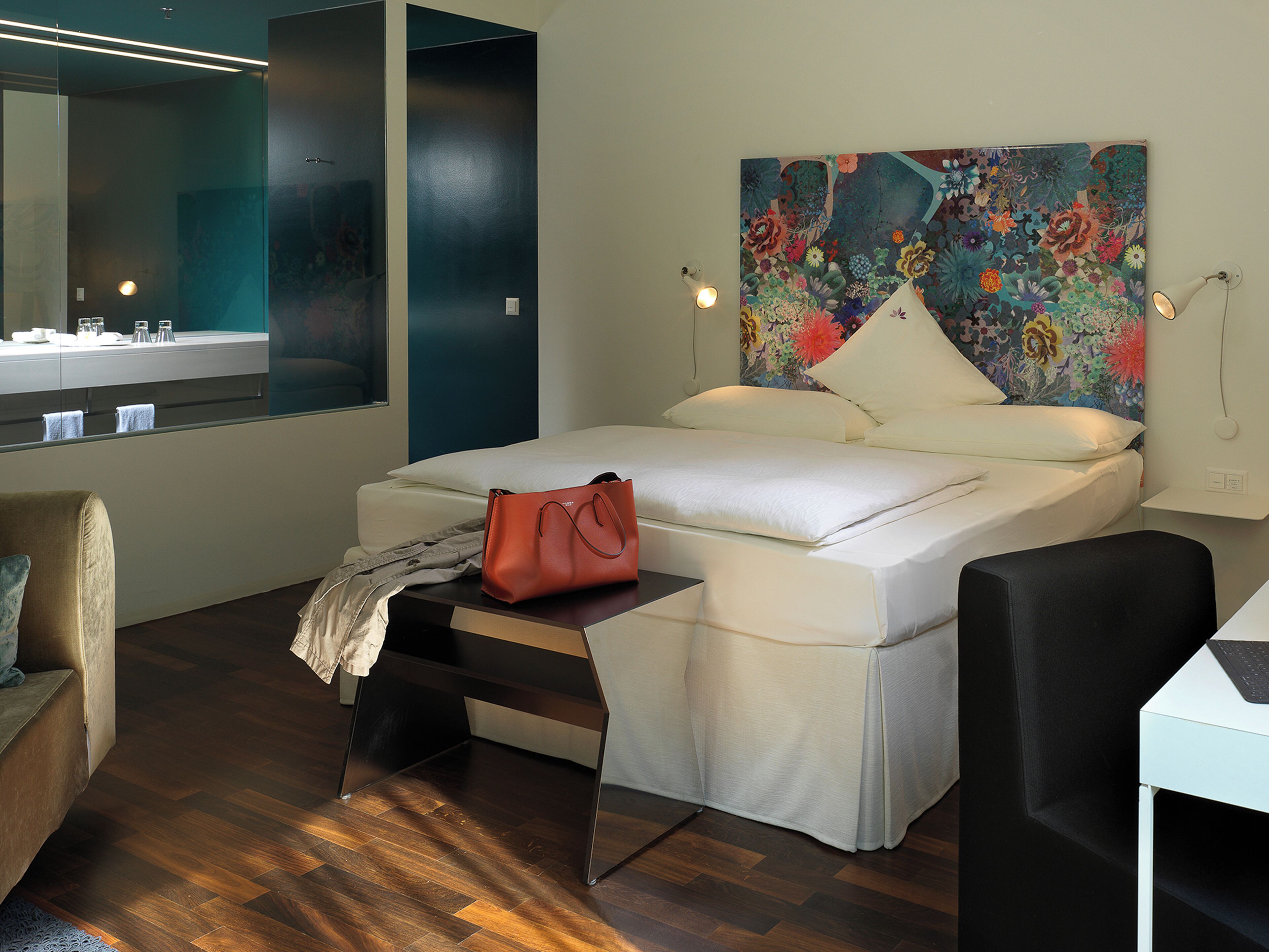 A modern bedroom with a bed, a desk, a chair, a mirror, and a large floral artwork on the wall.