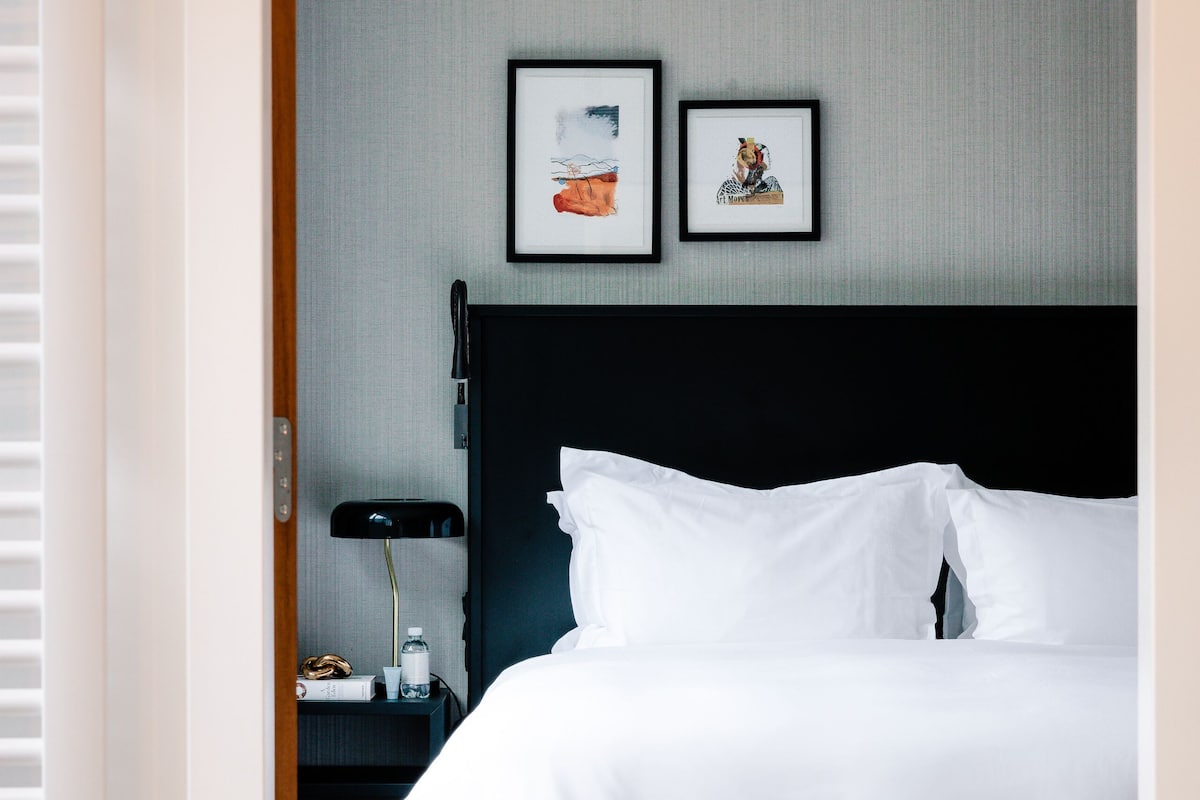 Habitación de hotel con una cama grande, una mesita de noche con lámpara y dos cuadros en la pared.