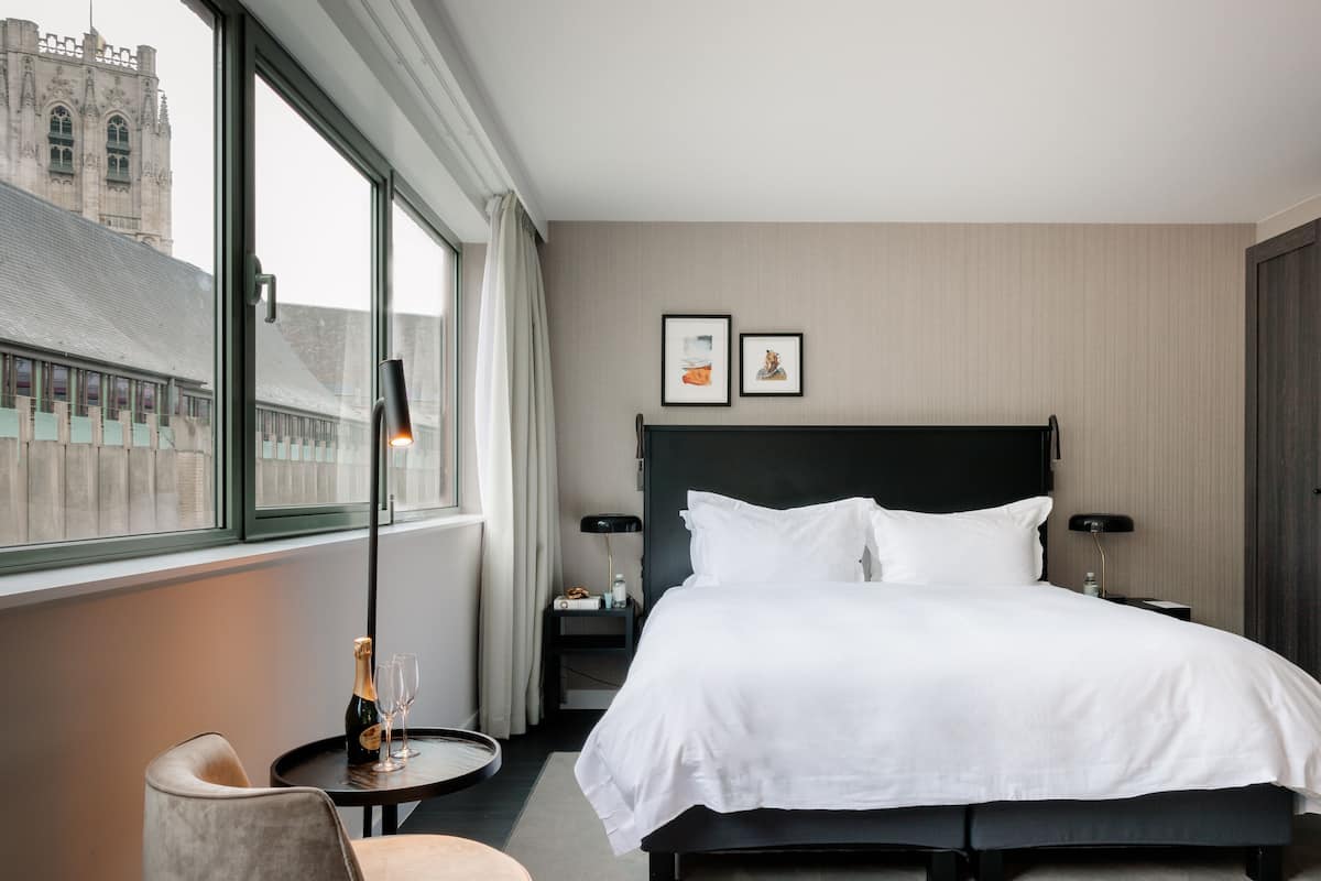Habitación de hotel con una cama grande, una mesita de noche, una silla, una pequeña mesa redonda con una botella y vasos, y vista a un edificio a través de la ventana.