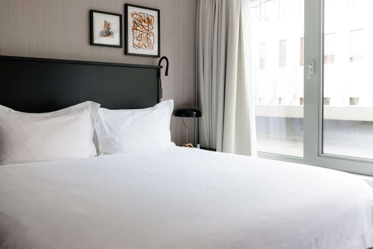 Una cama bien tendida con sábanas blancas, cabecera oscura, un cuadro enmarcado, una lámpara y vistas a través de una ventana.