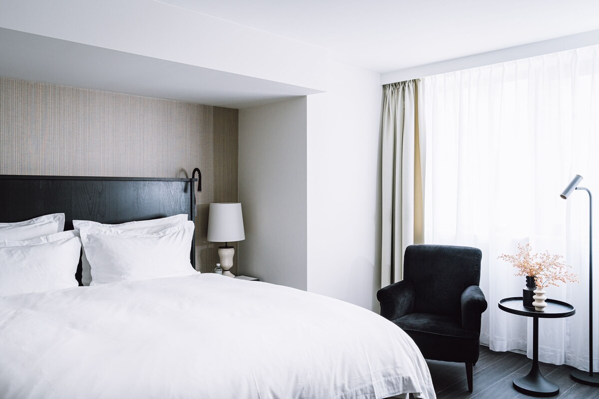 Ein Hotelzimmer mit einem großen Bett, einem Sessel, einem kleinen Tisch mit einer Vase und einer Lampe.