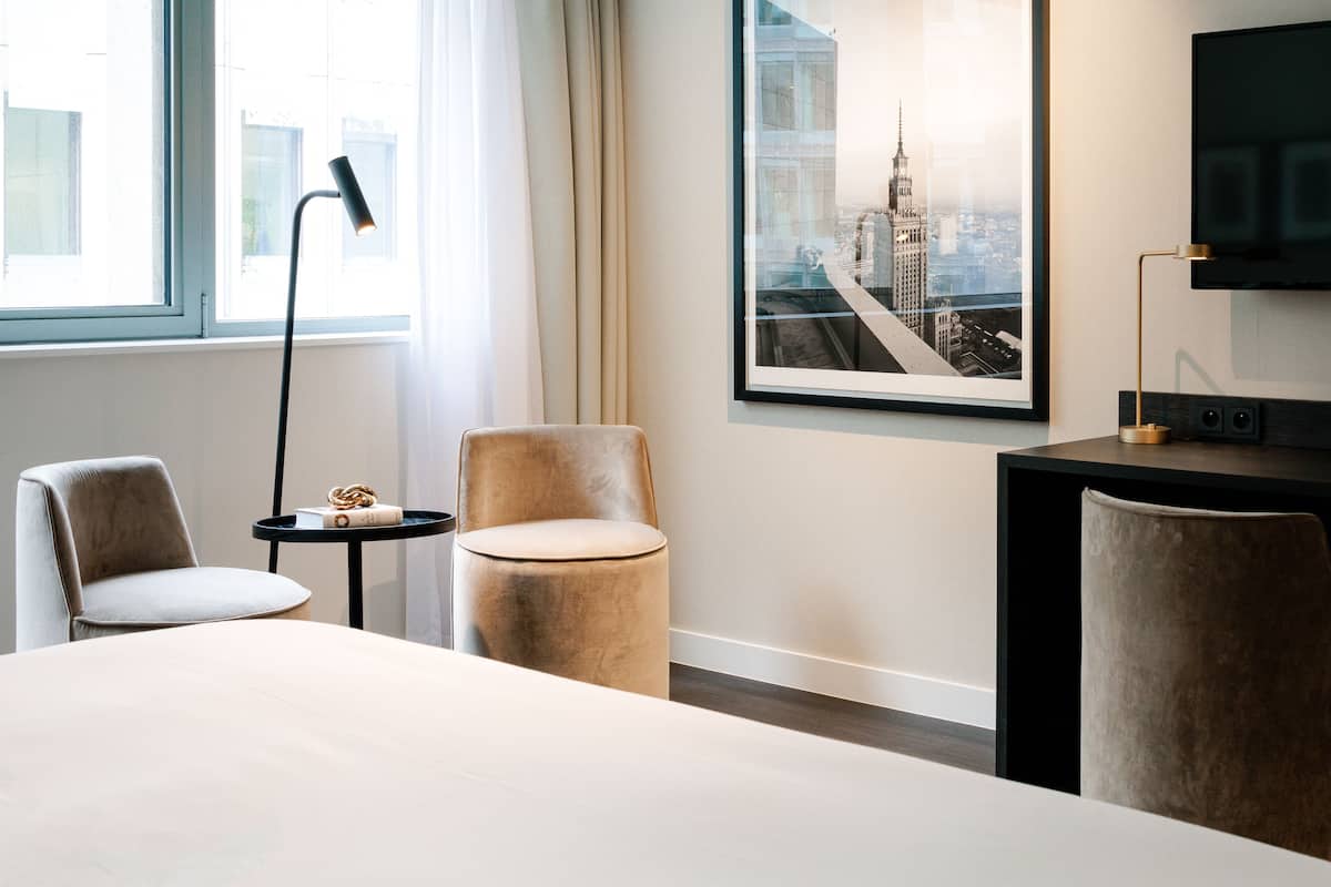 Habitación de hotel con cama, dos sillones, una mesita redonda, una lámpara, un cuadro enmarcado de un edificio y un televisor sobre un mueble.