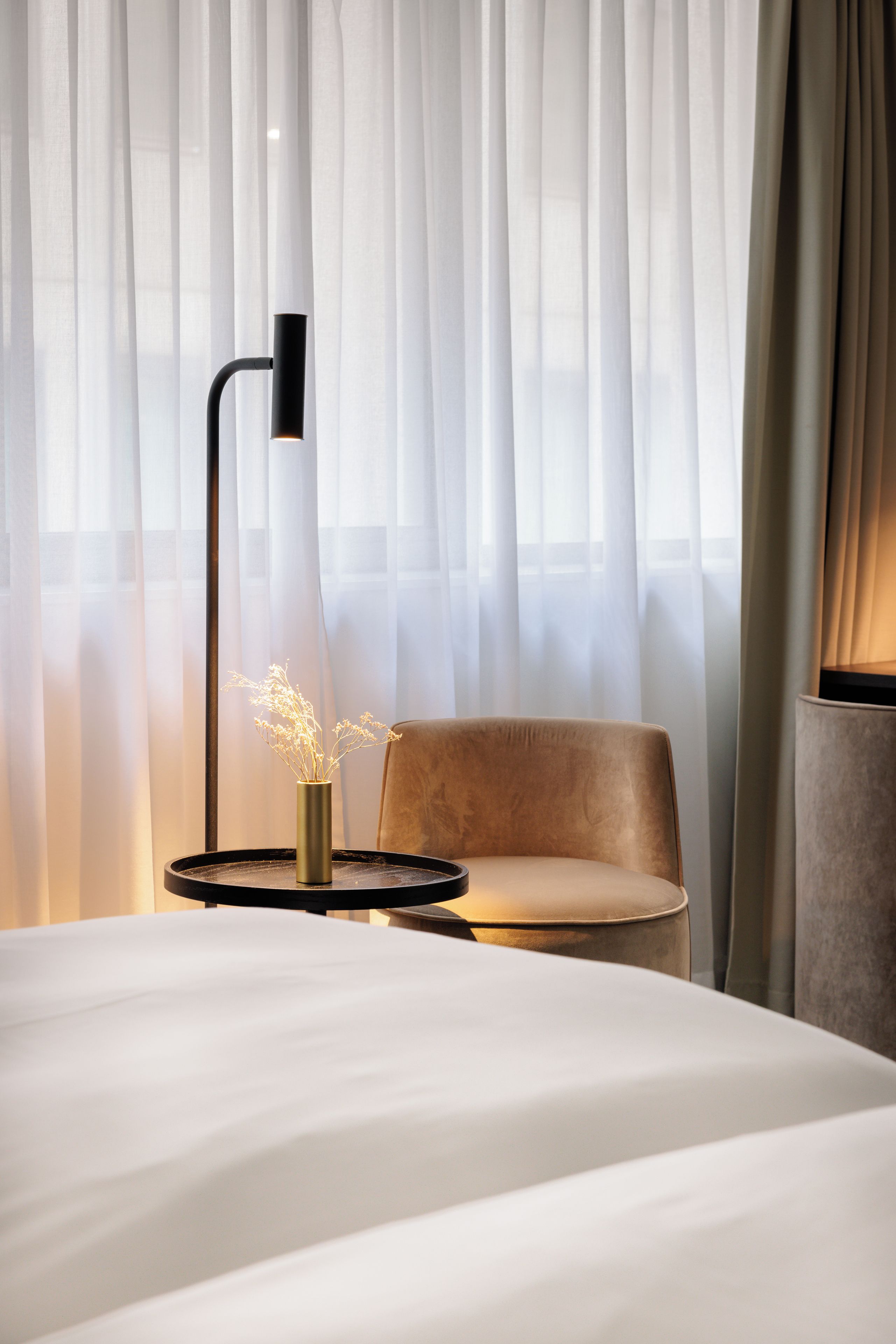 Une chambre d’hôtel avec un lit, une chaise, une table ornée d’un vase de fleurs et un lampadaire.