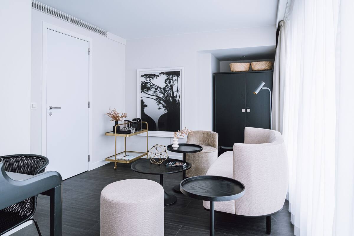 Una sala de estar moderna con un sofá, sillones, una mesa de centro y una mesita auxiliar. Se observa una estantería con objetos decorativos y un cuadro enmarcado en la pared.