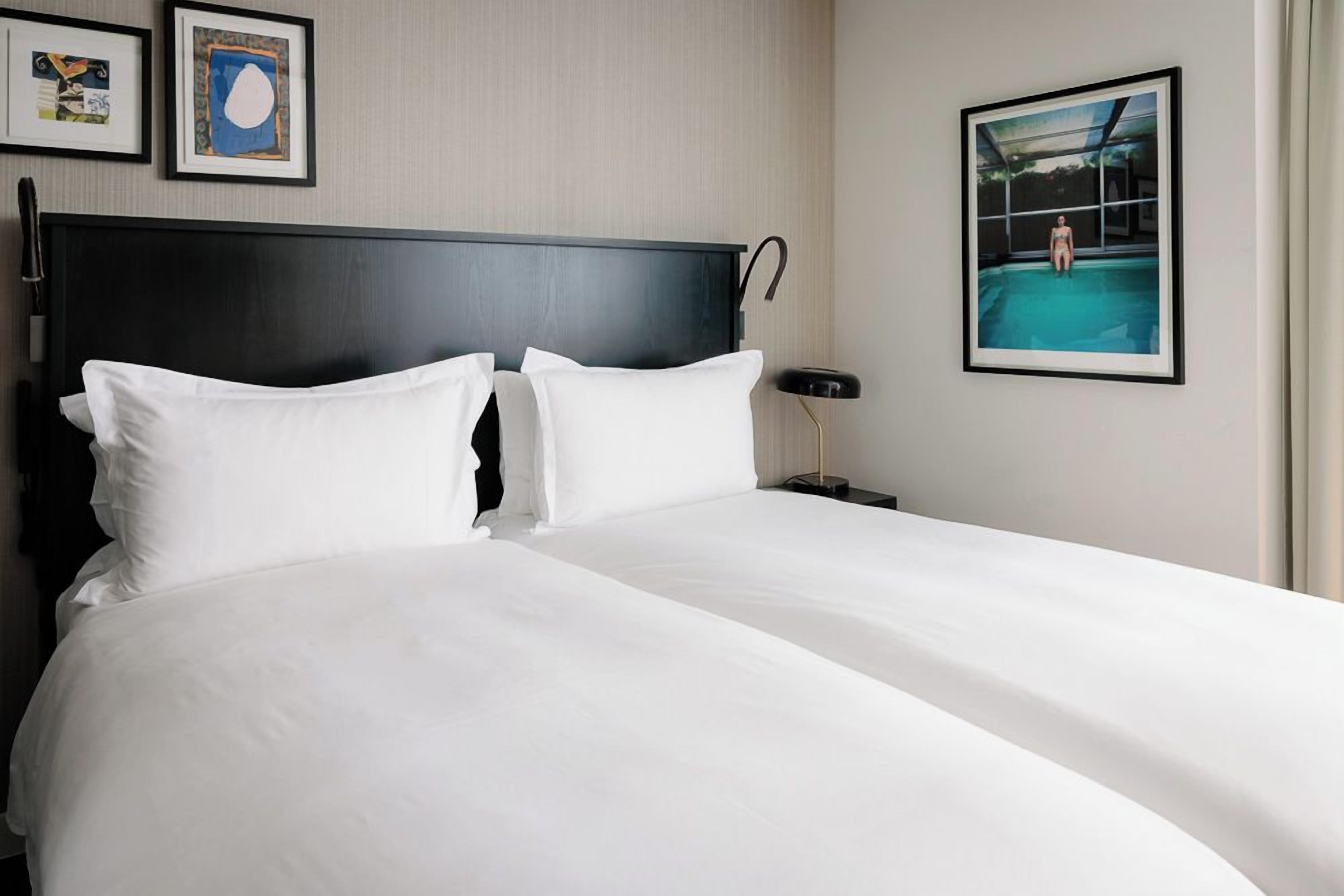 Habitación de hotel con una cama grande, dos almohadas, cabecero, mesita de noche, lámpara y cuadros en la pared.