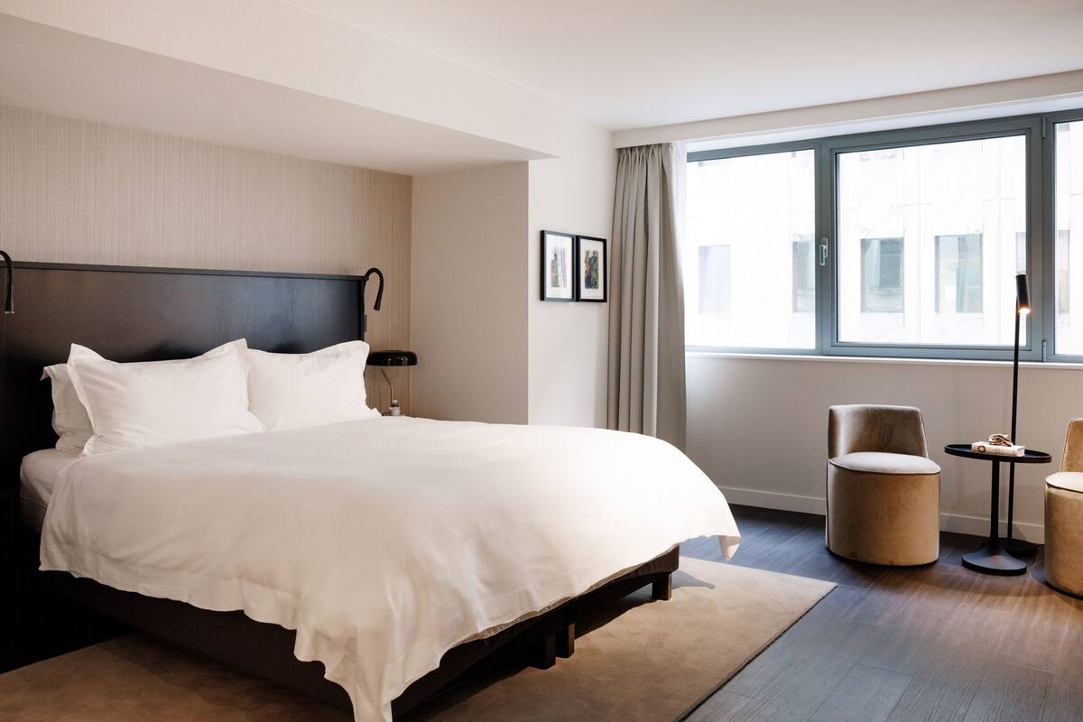 Una habitación de hotel moderna con una cama grande, una mesita de noche, una silla, una lámpara de pie y una ventana con cortinas.