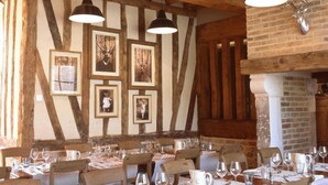 A rustic dining area with wooden beams, exposed brick walls, and framed pictures on the wall. Tables are set with wine glasses, water glasses, and cutlery. - Hôtel Le Grand Cerf & Spa Nuxe (Lyons-la-Foret)