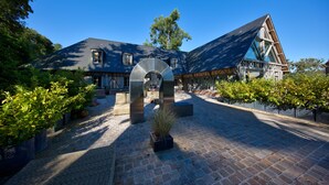 A building with a sloped roof, a central entrance with a large archway, and a brick pathway leading to it. - Hôtel Le Grand Cerf & Spa Nuxe (Lyons-la-Foret)