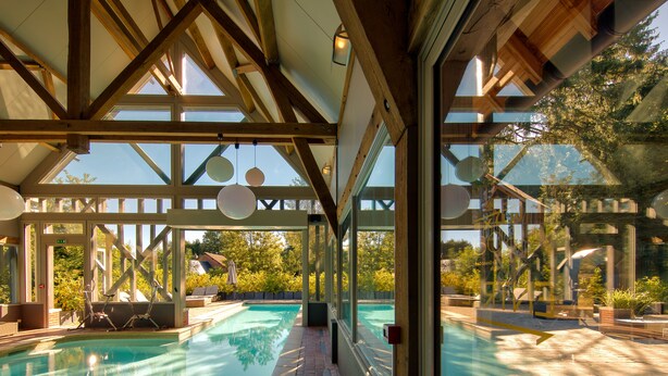 A swimming pool with a wooden ceiling and large windows, surrounded by potted plants and outdoor furniture. - Hôtel Le Grand Cerf & Spa Nuxe (Lyons-la-Foret)
