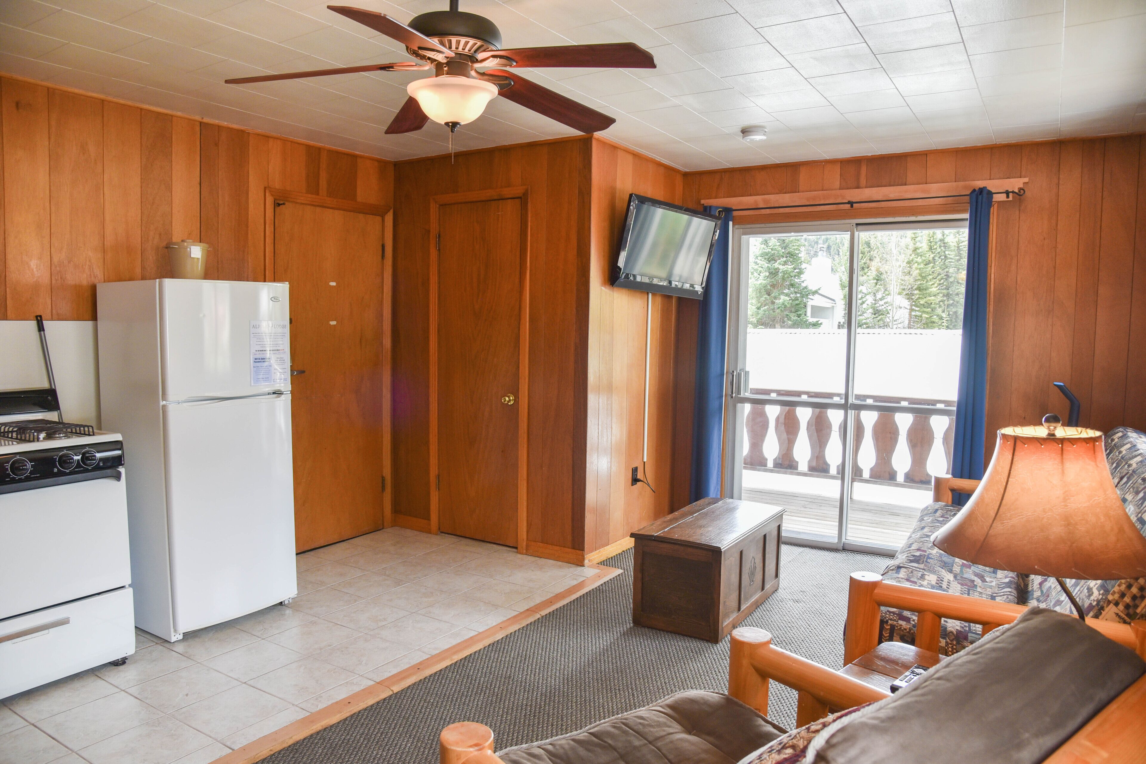 A compact kitchen with a refrigerator, stove, and ceiling fan. A television is mounted on the wall, and there is a sliding glass door leading to an outdoor area.