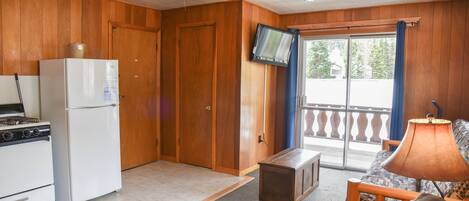 A compact kitchen with a refrigerator, stove, and ceiling fan. A television is mounted on the wall, and there is a sliding glass door leading to an outdoor area.