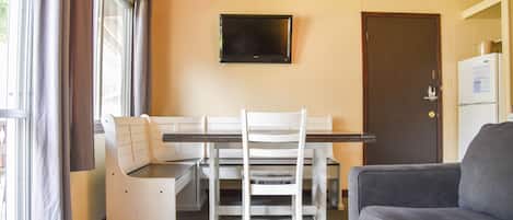Una habitación con una mesa de comedor, sillas, un televisor montado en la pared, una puerta y un refrigerador.