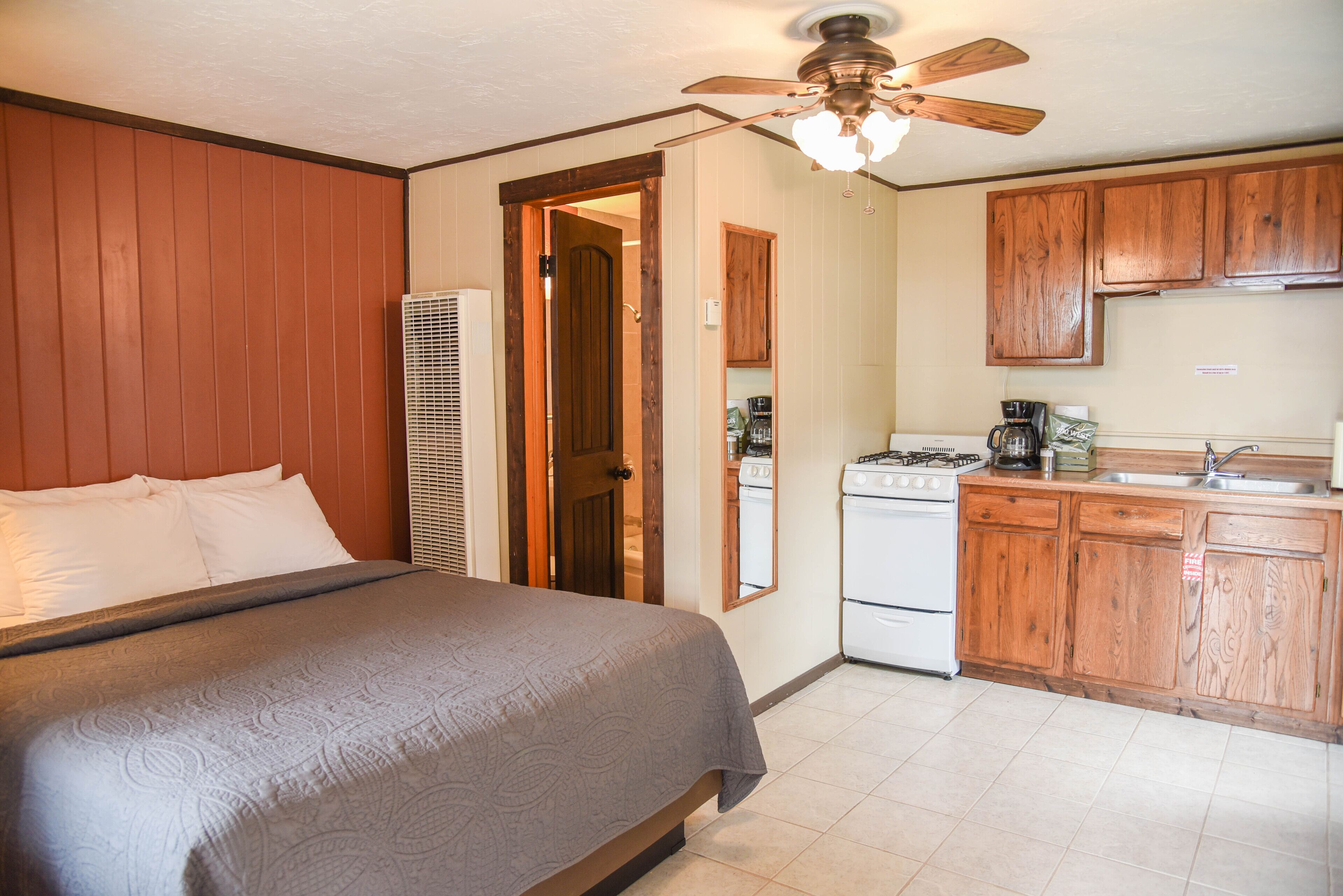 A bedroom with a bed, a ceiling fan, and a kitchenette with wooden cabinets and a stove.