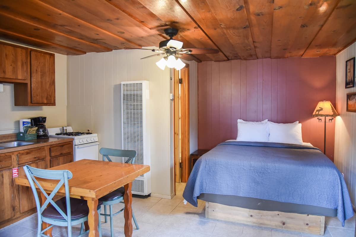 A room with a bed, a wooden ceiling, a dining table with chairs, a kitchen area with cabinets and appliances, and a wall-mounted lamp.